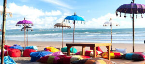 A beach scene with colorful bean bags and cushions arranged on the sand under vibrant parasols. The parasols are adorned with decorative fringes, and a wooden table is placed among the seating. The ocean waves are gently rolling in under a bright blue sky with scattered white clouds.