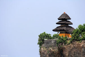 A traditional thatched-roof pavilion perched on the edge of a steep cliff, surrounded by lush greenery. The pavilion has multiple tiers and overlooks a vast, clear sky, suggesting an elevated and serene location.