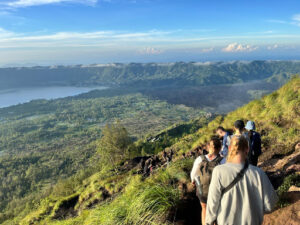 A group of hikers walking along a mountain trail with a scenic view of a lush, green valley below. The valley includes a lake and expansive forested areas, with mountains in the distance under a clear blue sky. The hikers are dressed casually and appear to be enjoying the panoramic landscape