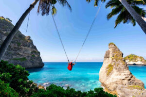 A person wearing a red dress is swinging on a swing suspended between two palm trees, overlooking a stunning turquoise sea with rocky cliffs and lush greenery in the background. The scene is bright and sunny, showcasing a beautiful tropical paradise