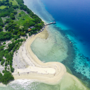 An aerial view of a tropical island with a sandy beach, lush greenery, and crystal-clear blue water. A small dock is visible on the shore, and a boat is anchored nearby. The water is shallow and turquoise near the shore, and it gradually deepens as it moves away from the island.