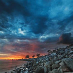  A stunning sunset over a calm lake. The sky is ablaze with vibrant colors of orange, pink, and purple, with dramatic dark clouds. A small group of trees stands on a rocky shoreline